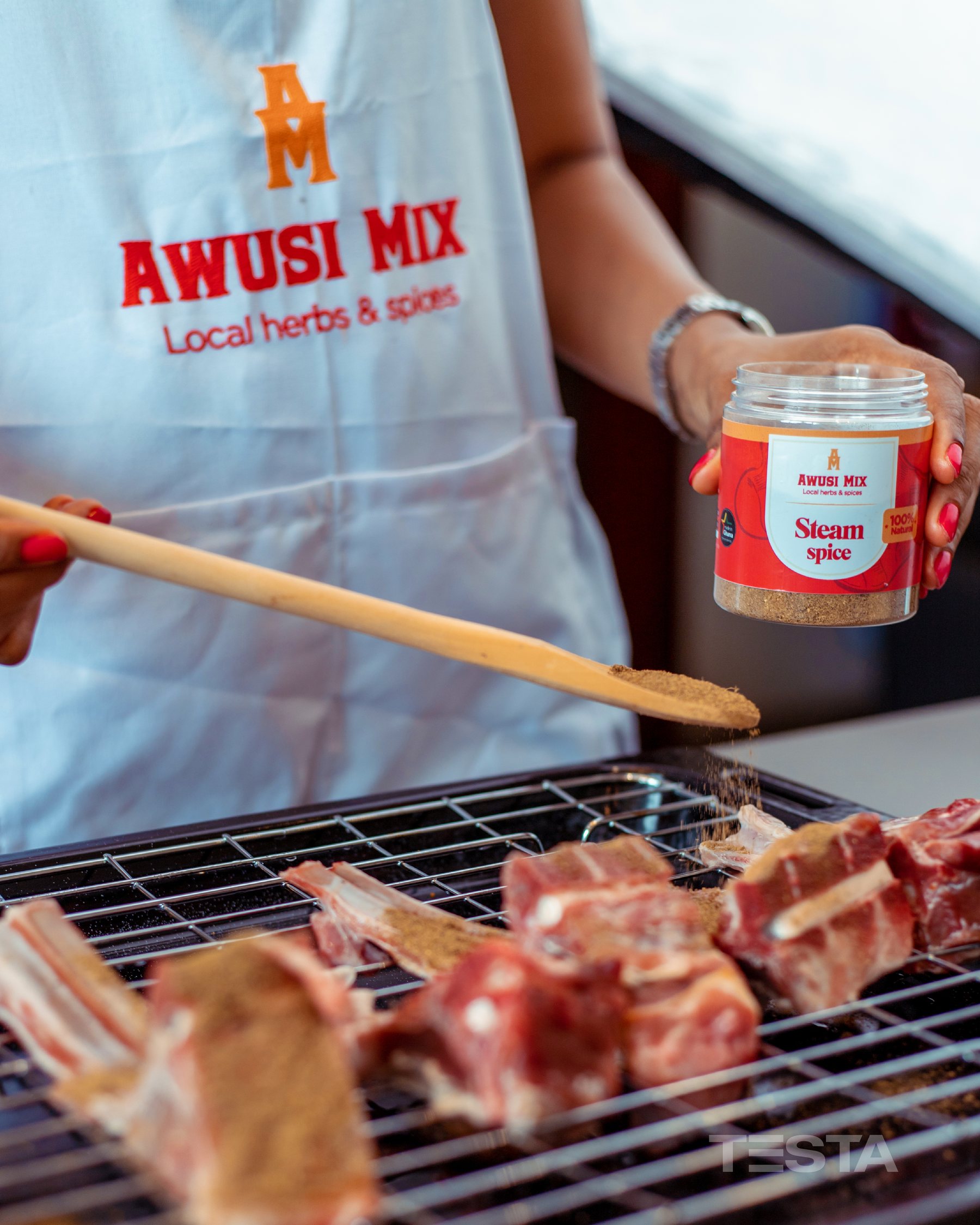 A cook in an Awusi Mix apron seasoning grilling lamb chops with a jar of Steam Spice